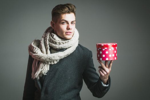 Handsome sexy man in white knitted winter scarf and jacket has fashionable gift. Young stylish guy holds red spotted present box for valentines or christmas holidays on grey background.