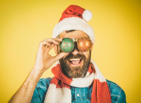 Christmas hipster happy smile in santa hat and scarf. Man cover eyes with xmas balls on yellow background. Holiday decorations.