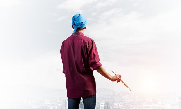 Young male painter artist holding paintbrush. Back view of painter in shirt and bandana on background cloudy sky and cityscape. Creative hobby and artistic occupation. Art school courses concept.