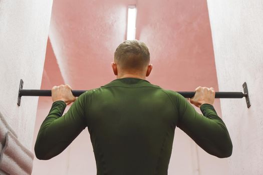 Young sports guy in a green thermal suit pulls himself up on the horizontal bar in the gym.