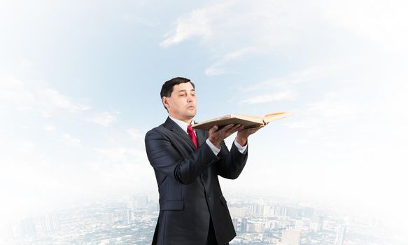 Serious businessman holding open old book. Adult man in business suit standing on cloudy cityscape background. Lawyer reading big legal regulation book. Professional business accounting and consulting