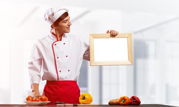 Young male chef standing near cooking table and holding blank white board. Handsome chef in white and red uniform in light kitchen interior. Cooking classes advertising. Professional catering service