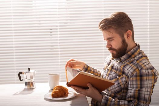 Side view of pensive young bearded hipster man reading book and having dinner with croissant and cup of coffee on background of blinds.