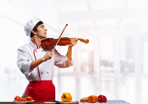 Young male chef with violin standing near cooking table with vegetables. Handsome chef in white hat and red apron in light kitchen interior. Creative culinary performance. High cooking skills concept