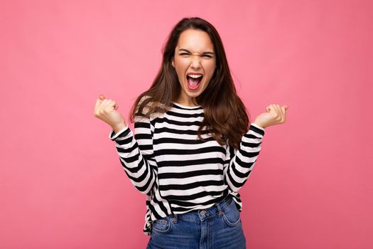 Portrait of young positive happy beautiful brunette woman with sincere emotions wearing casual striped pullover isolated on pink background with empty space and celebrating victory saying yeah.