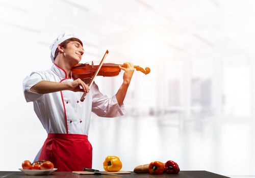 Young male chef with violin standing near cooking table with vegetables. Handsome chef in white hat and red apron in light kitchen interior. Creative culinary performance. High cooking skills concept