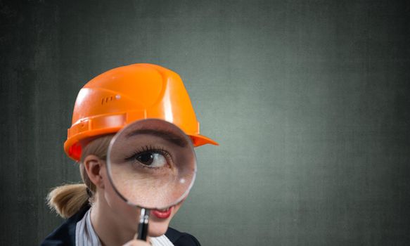Professional appraiser looking through magnifying glass. Woman civil engineer in safety helmet checking with magnifier on wall background. Technical condition assessment, consultancy and expertise