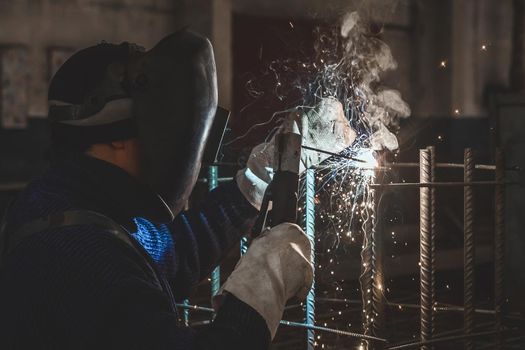 A masked working man is doing welding work on metal structures in a factory or industrial enterprise.