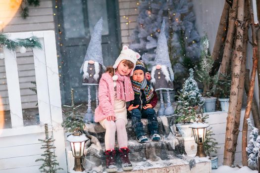 Happy little kids sitting on the porch of the Christmas decorated house, snowing outdoor. Happy New Year and Merry Christmas. Magic winter