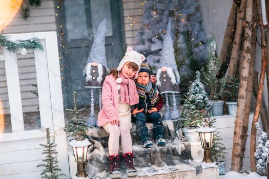Happy little kids sitting on the porch of the Christmas decorated house, snowing outdoor. Happy New Year and Merry Christmas. Magic winter