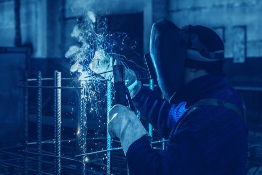 Working man is doing welding work on metal structures in a factory or industrial enterprise.