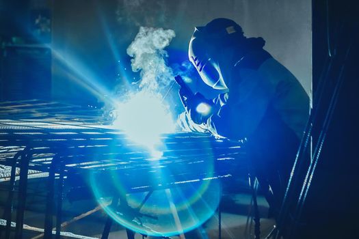 Working man is doing welding work on metal structures in a factory or industrial enterprise.