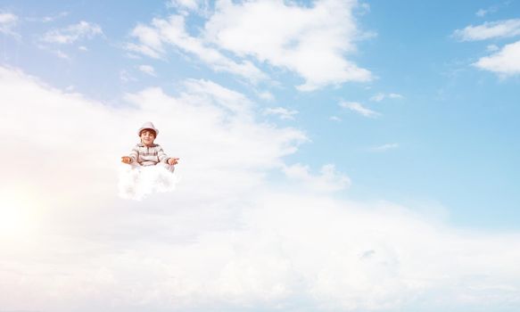 Young little boy keeping eyes closed and looking concentrated while meditating on clouds in the air with cloudy skyscape on background.