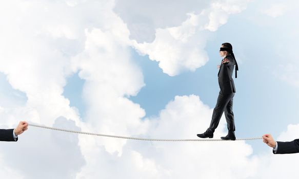 Businessman with blindfolder on eyes walking on rope over sky background