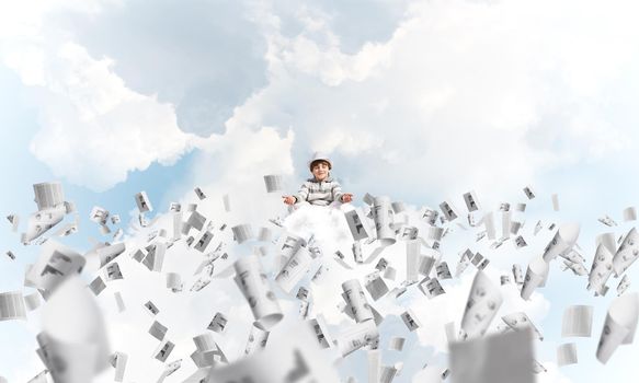Young little boy keeping eyes closed and looking concentrated while meditating among flying papers in the air with cloudy skyscape on background.