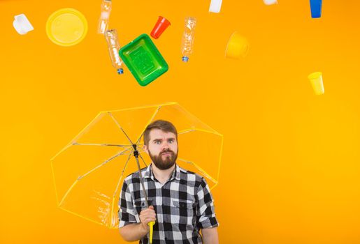 Plastic recycling problem, ecology and environmental disaster concept - Angry man hiding from garbage under an umbrella on yellow background.