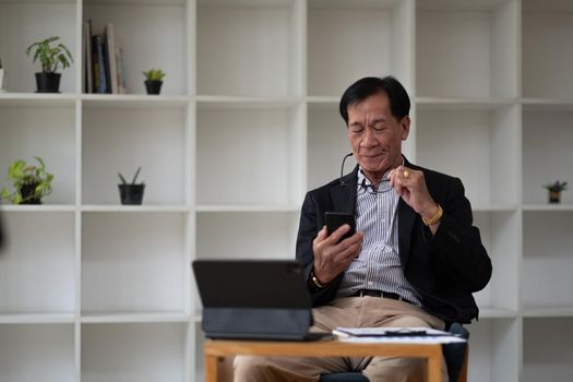 Asian old elder senior man on mobile smart phone and digital tablet on wooden desk.