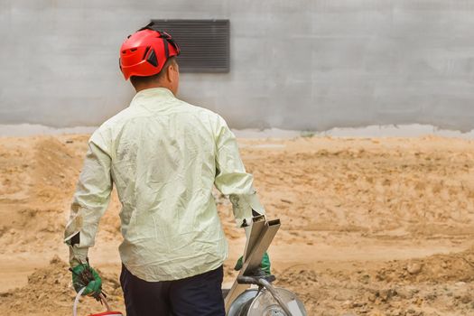 Industrial worker in a red helmet walks with a working tool against the background of a sand and concrete building house at a construction site.