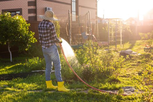 Caucasian woman gardener in work clothes watering the beds in her vegetable garden on sunny warm summer day. Concept of working in the garden
