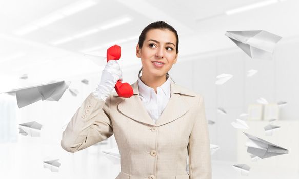 Smiling young woman holding retro red phone in office with flying paper planes. Call center operator in business suit with landline phone. Hotline telemarketing. Business assistance and consultation.
