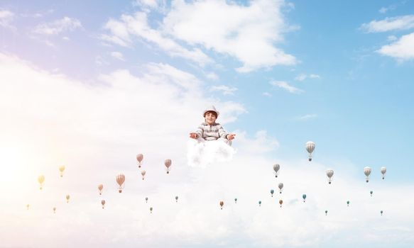 Young little boy keeping eyes closed and looking concentrated while meditating among flying balloons in the air with cloudy skyscape on background.