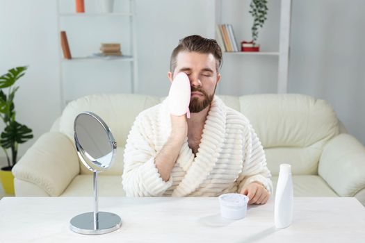 Bearded man look in mirror and clean scrub face with sponge, happy millennial male metrosexual do beauty procedures at home, take care of healthy skin, skincare concept.