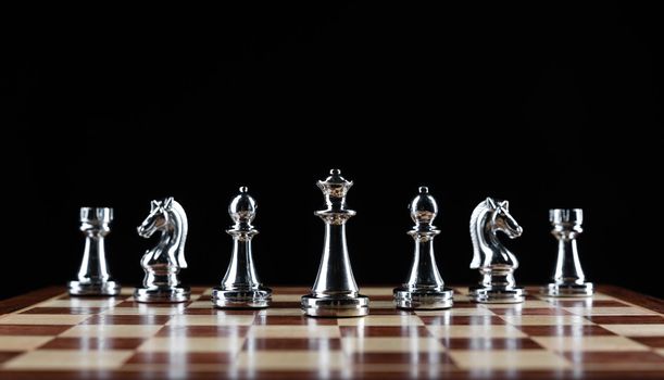 Shiny steel chess figures standing on wooden chessboard. Intellectual duel and tactical battle symbol. Strategy planning and corporate leadership concept. Silver metal chess pieces on black background