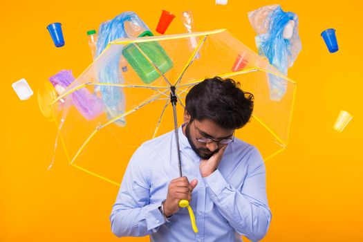 Plastic recycling problem and environmental disaster concept - Indian man hiding from garbage under an umbrella on yellow background.