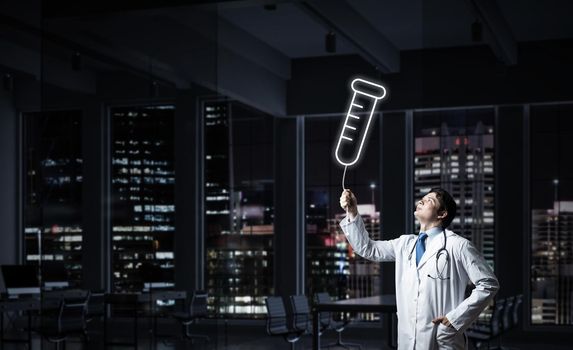 Horizontal shot of young confident doctor in white medical uniform interracting with glowing vial symbol whie standing against night cityscape view on background.