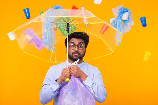 World Environment Day, plastic recycling problem and environmental disaster concept - Sad Indian man hiding from garbage under an umbrella on yellow background.