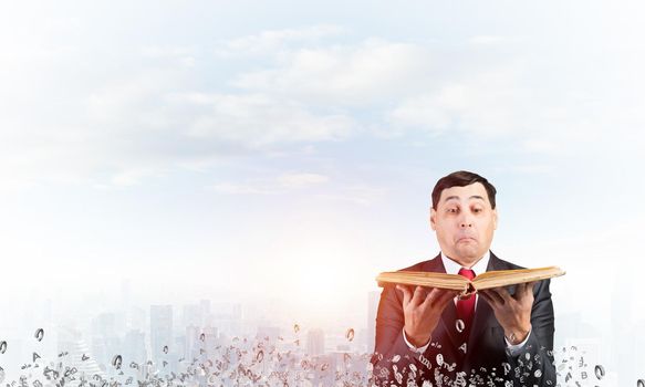 Surprised businessman looking into open old book. Startled man in business suit and tie standing on downtown background with flying various letters. Business education and knowledges concept.