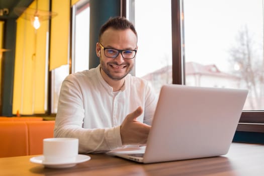 Portrait of a young handsome businessman in a white shirt, glasses, with a beard sits at a table in a laptop over a cup of coffee and smiles.