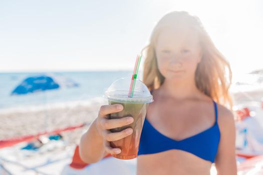 A young girl with blonde hair of European appearance, a teenager holds a colored cold non-alcoholic cocktail in her hand against the background of the sea beach.