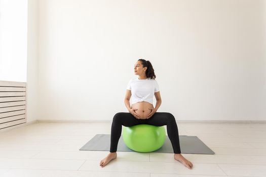 A young pregnant woman doing relaxation exercise using a fitness ball while sitting on a mat and holding her tummy