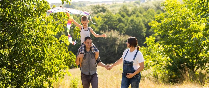 Happy family with pregnant wife fly a kite together in summer field