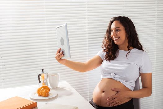 Happy pregnant young beautiful woman talking to mom using video call during morning breakfast. Communication and positive attitude during pregnancy