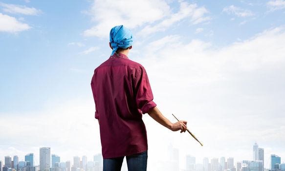 Young male painter artist holding paintbrush. Back view of painter in shirt and bandana on background cloudy sky and cityscape. Creative hobby and artistic occupation. Art school courses concept.