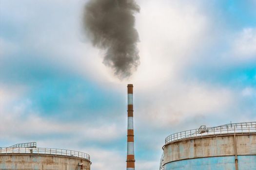 Problems of environmental pollution and ecology concept. Gray smoke comes from a chimney of an industrial plant or thermal power plant next storage tanks against the background of the sky.