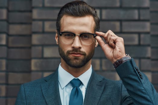 Smart and successful. Confident businessman in formal wear adjusting his eyeglasses and looking at camera while standing against brick wall. Business concept. Stylish people