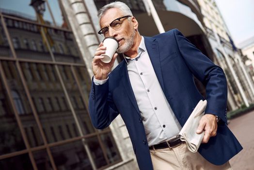 Handsome senior man in smart casual wear carrying disposable cup while walking through the city street