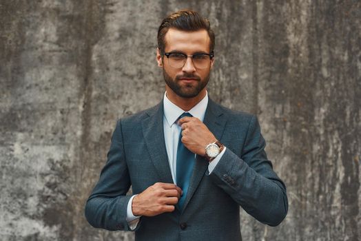 Perfect look. Confident businessman in full suit adjusting his tie and looking at camera while standing against grey stone wall outdoors. Business concept. Stylish people
