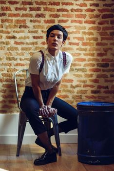 Beautiful young woman, model of fashion, with very short haircut, looking at camera sitting on chair, with a brick wall in the background. Beauty and hairstyles concept.