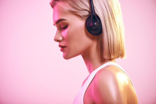 Music waves. Side view of beautiful blonde woman in headphones listening music while standing against pink background. Sport. Music concept