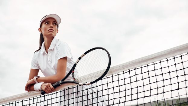 Portrait of sportswoman in white uniform and cap with tennis racket posing near tennis net on court. Horizontal shot