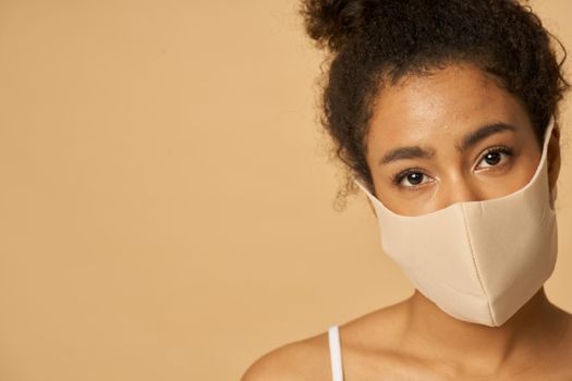 Close up portrait of beautiful young mixed race woman wearing protective facial mask looking at camera while posing isolated over beige background. Safety, pandemic concept