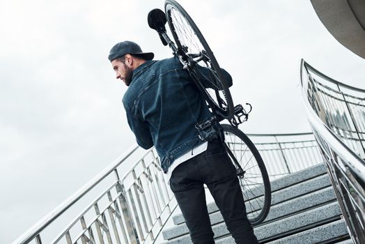 Outdoors leisure. Young stylish man walking up the stairs holding bicycle looking aside curious