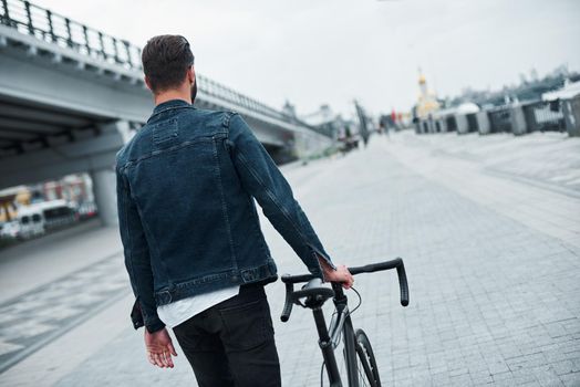 Outdoors leisure. Young stylish man walking on city street with bicycle back view