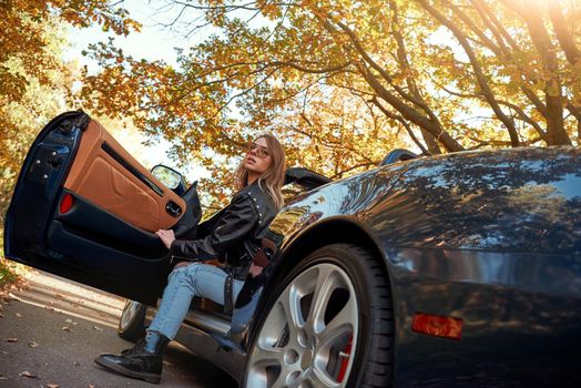 Beautiful blonde girl in the roofless car. Autumn season, leather coat and jeans, stylish sunglasses on head. Rear view