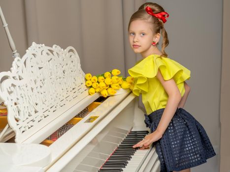 A sweet schoolgirl girl sits beside a white grand piano on which lies a bouquet of flowers. The concept of studying music, spring mood, happy people.