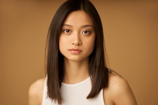 Face closeup of beautiful mixed race young woman with perfect skin looking at camera while standing isolated over brown background. Female beauty portrait